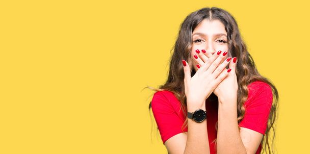 Junge schöne Frau in lässigem T-Shirt schockiert Mund mit Händen für Fehler bedecken. Geheimes Konzept. - Foto, Bild