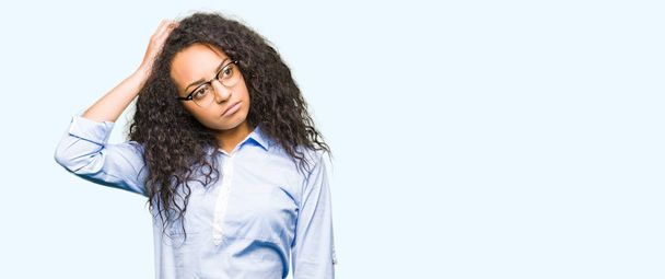 Young beautiful business girl with curly hair wearing glasses confuse and wonder about question. Uncertain with doubt, thinking with hand on head. Pensive concept. - Photo, Image
