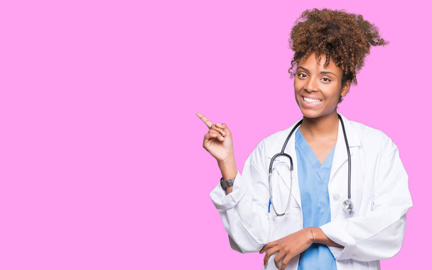 Nuori afrikkalainen amerikkalainen lääkäri nainen yli eristetty tausta iso hymy kasvoillaan, osoittaen käsi ja sormi sivulle katsoen kameraa
. - Valokuva, kuva