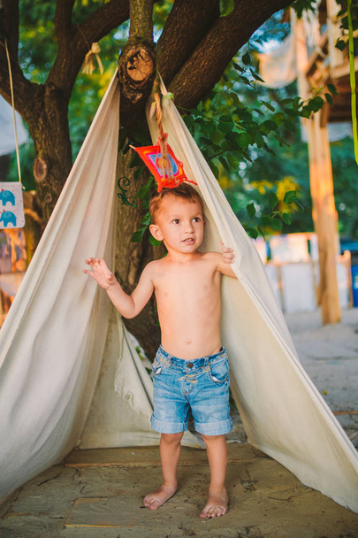 theme summer vacation, little boy, Caucasian child playing in wooded area in park on playground in yard. kid in Tipi wigwam tent in summer. Explore and play in nature on summer time - Photo, Image