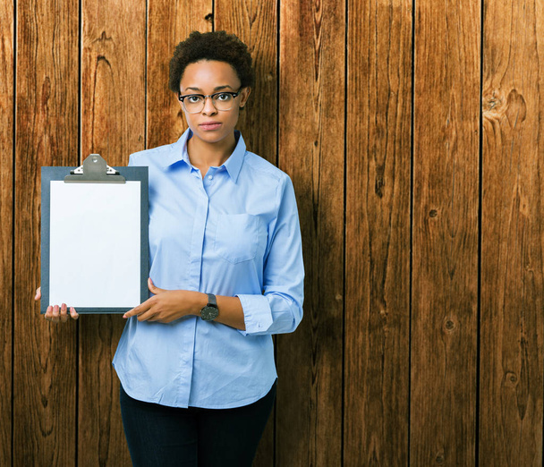 junge afrikanisch-amerikanische Geschäftsfrau hält Klemmbrett über isolierten Hintergrund mit einem selbstbewussten Gesichtsausdruck auf intelligentem Gesichtsdenken ernst - Foto, Bild