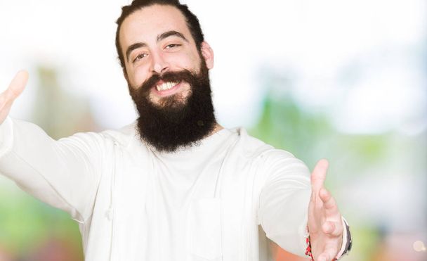 Young man with long hair and beard wearing sporty sweatshirt looking at the camera smiling with open arms for hug. Cheerful expression embracing happiness. - 写真・画像