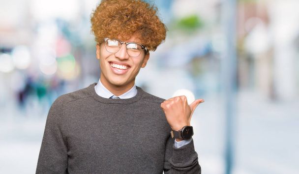 Jonge knappe Business man met Afro haar dragen bril glimlachend met gelukkig gezicht kijken en wijzend naar de zijkant met duim omhoog. - Foto, afbeelding