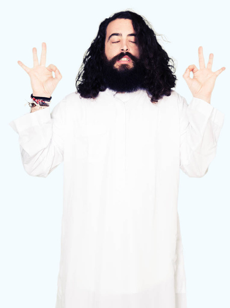 Hombre vistiendo traje de Jesucristo relajarse y sonreír con los ojos cerrados haciendo gesto de meditación con los dedos. Concepto de yoga
. - Foto, Imagen