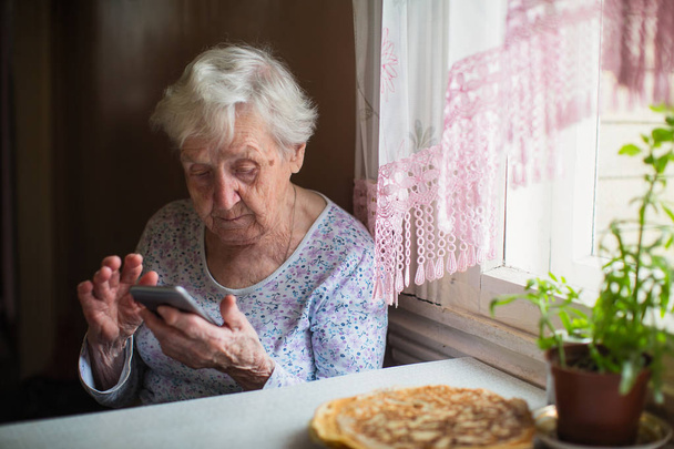 Yaşlı bir kadın evde elinde bir akıllı telefon ile oturur. - Fotoğraf, Görsel