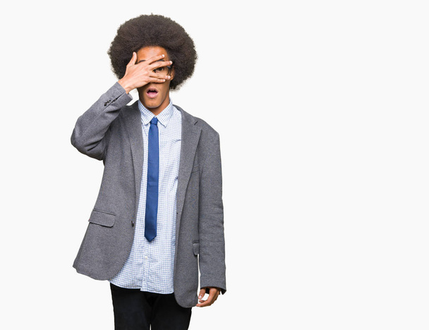 Young african american business man with afro hair wearing glasses peeking in shock covering face and eyes with hand, looking through fingers with embarrassed expression. - Foto, Imagen