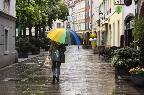 Μια γυναίκα με ομπρέλα από ουράνιο τόξο περπατάει στην πόλη στη βροχή. Concept φθινόπωρο, κακοκαιρία - Φωτογραφία, εικόνα