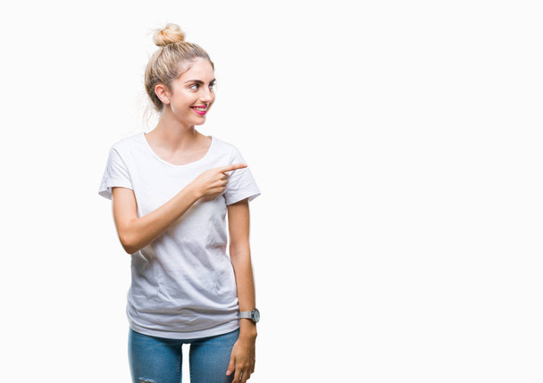 Young beautiful blonde woman wearing white t-shirt over isolated background cheerful with a smile of face pointing with hand and finger up to the side with happy and natural expression on face looking at the camera. - Fotografie, Obrázek