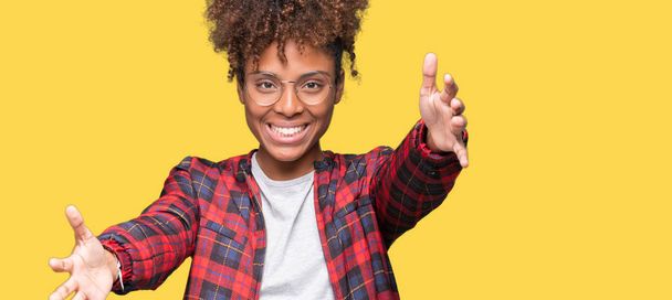 Beautiful young african american woman wearing glasses over isolated background looking at the camera smiling with open arms for hug. Cheerful expression embracing happiness. - Фото, изображение