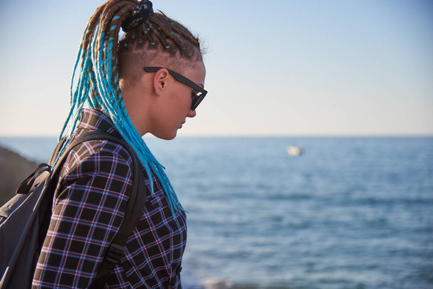 Κορίτσι με μπλε ράμες και σκούρα γυαλιά στη θάλασσα, φωτισμένο από τον απογευματινό ήλιο. - Φωτογραφία, εικόνα