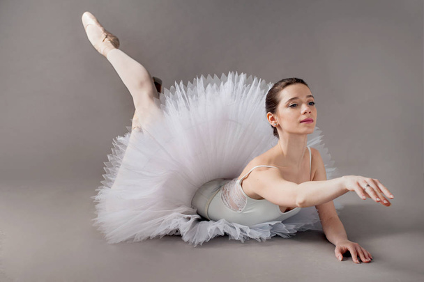 Una bailarina elegante joven vestida con un tutú blanco, demuestra habilidades de baile posando mientras está sentada en el piso del estudio
. - Foto, imagen