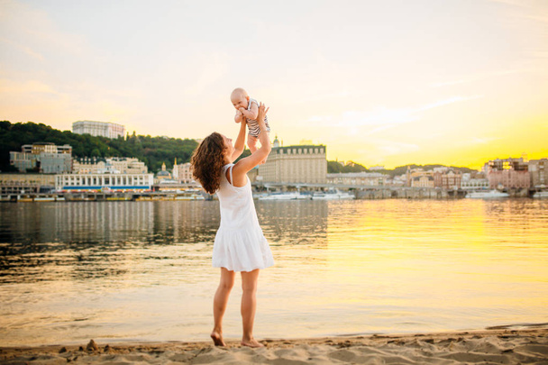 夕日と水に対して子供を回す母親。幸せなママと赤ちゃんビーチで遊ぶ。息子を抱く若い女性 - 写真・画像