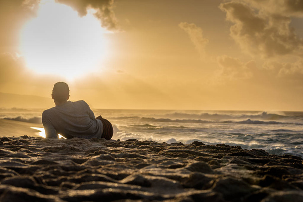 Silueta de un joven caucásico sentado en Sunset Beach en Hawaii mirando hacia el atardecer sobre el océano
 - Foto, imagen