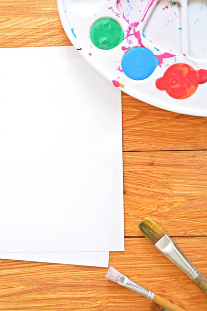 ein Blatt Papier mit Farben in einer Farbpalette und vielen Pinseln sowie Pinselwasser auf einem Holzgrund - Foto, Bild