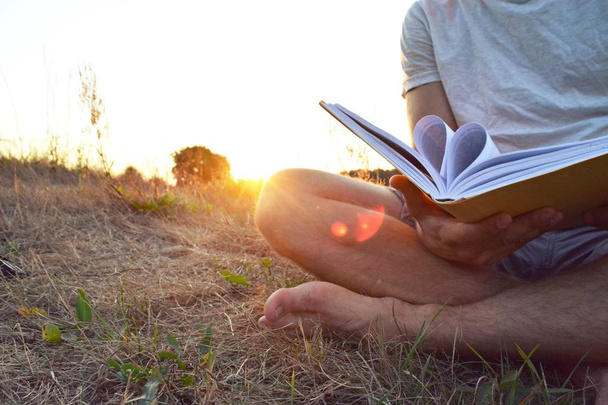 Close-up - hombre leyendo un libro en el campo sin zapatos y pantalones cortos mientras el sol se pone en el fondo cerca de un bosque - concepto para el amor de la lectura
  - Foto, Imagen