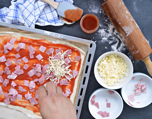 Close-up - Man is filling a pizza by himself with ham, salami and cheese - baking sheet full of pizza dough with ingredients next to it  - Foto, Bild