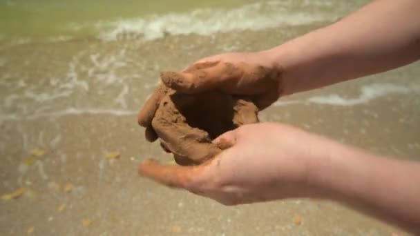 Κοντινό πλάνο των χεριών που κάνουν πηλό σε μια παραλία θάλασσα, άμμο και κύματα ως φόντο, το καλοκαίρι και το ταξίδι έννοια. - Πλάνα, βίντεο