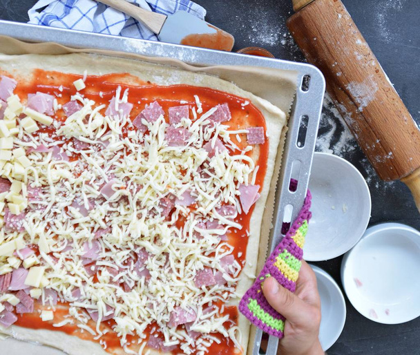 Close-up - El hombre está llenando una pizza por sí mismo con jamón, salami y queso - bandeja para hornear llena de masa de pizza con ingredientes a su lado
  - Foto, Imagen