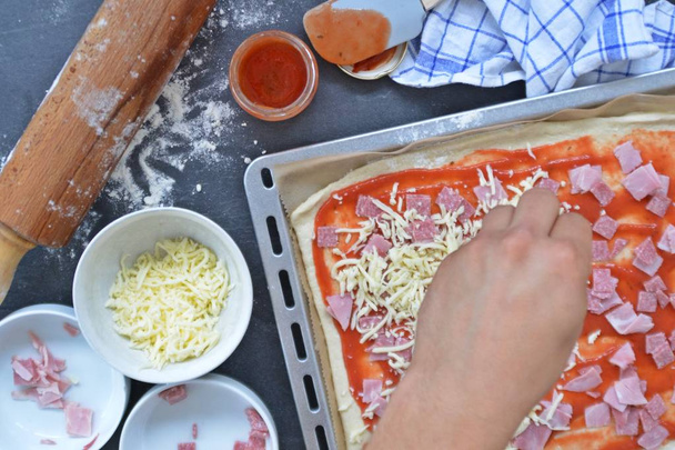 Close-up - El hombre está llenando una pizza por sí mismo con jamón, salami y queso - bandeja para hornear llena de masa de pizza con ingredientes a su lado
  - Foto, Imagen