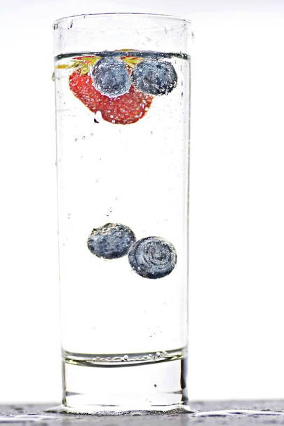 cair mirtilos e morangos em um copo cheio de água cintilante - close-up sobre o vidro isolado
 - Foto, Imagem