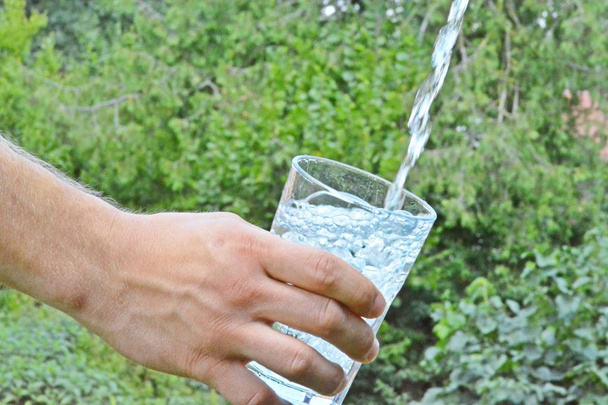 Vers en schoon water stroomt in een glas voor een groene zomerse achtergrond en wordt gehouden door de hand van een man  - Foto, afbeelding