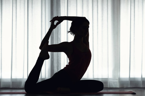 Silhouette of sporty young woman practicing yoga indoors - Foto, immagini