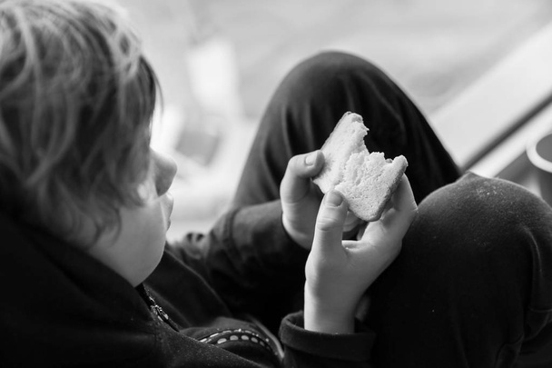 屋内の窓枠に座っているパンを持つホームレスの小さな男の子 - 写真・画像