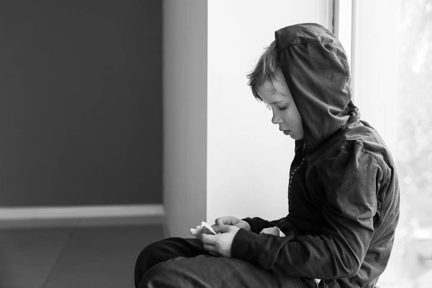 Homeless little boy with bread sitting on window sill indoors - Photo, Image