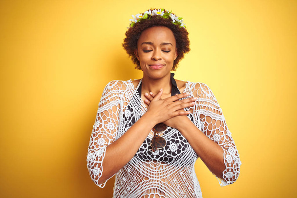 Młoda afroamerykańska kobieta z Afro włosy noszą kwiaty korony nad żółtym białym tle uśmiechnięty z rąk na klatce piersiowej z zamkniętymi oczami i wdzięczny gest na twarzy. Koncepcja zdrowia. - Zdjęcie, obraz