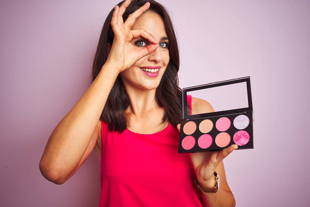 Young beautiful woman applying make up over pink isolated background with happy face smiling doing ok sign with hand on eye looking through fingers - Photo, Image