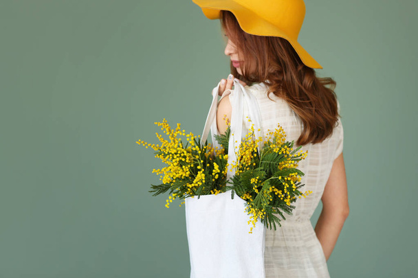 色の背景上のミモザの花の花束と美しい若い女性保持袋 - 写真・画像