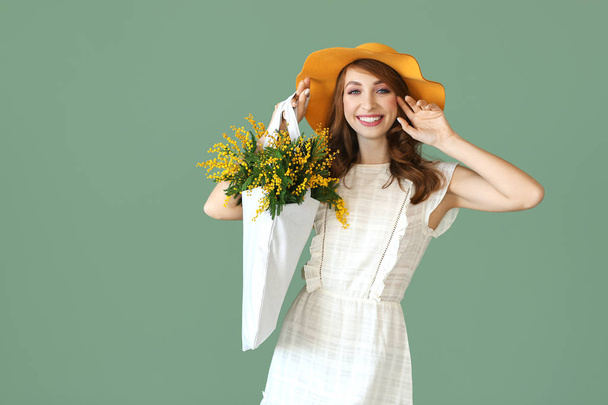 Beautiful young woman holding bag with bouquet of mimosa flowers on color background - Photo, Image