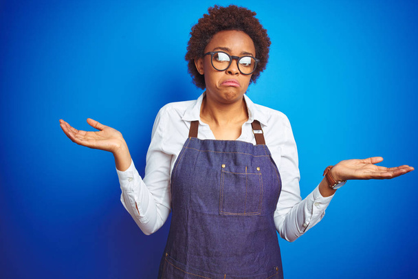Young african american woman shop owner wearing business apron over blue background clueless and confused expression with arms and hands raised. Doubt concept. - Photo, Image