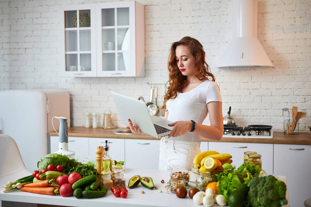 Jeune belle femme utilisant un cahier tout en cuisinant dans la cuisine moderne. Aliments sains et régime concept. Perdre du poids
 - Photo, image