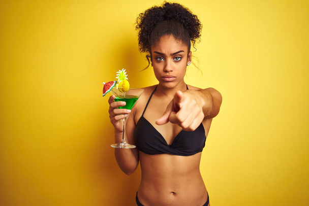 Femme afro en vacances portant un bikini buvant un cocktail sur fond jaune isolé pointant du doigt la caméra et vers vous, signe de la main, geste positif et confiant de l'avant
 - Photo, image
