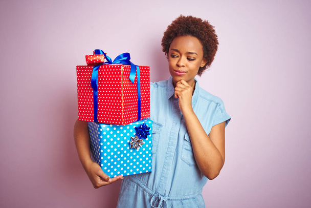 Afro-Amerikan kadın pembe izole arka plan üzerinde doğum günü hediyeleri tutan ciddi yüz soru hakkında düşünme, çok karışık fikir - Fotoğraf, Görsel