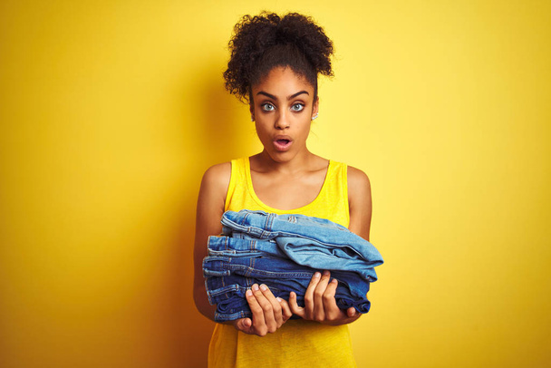 Jonge Afro-Amerikaanse vrouw met stapel jeans over geïsoleerde gele achtergrond bang in shock met een verrassings gezicht, bang en opgewonden met angst expressie - Foto, afbeelding