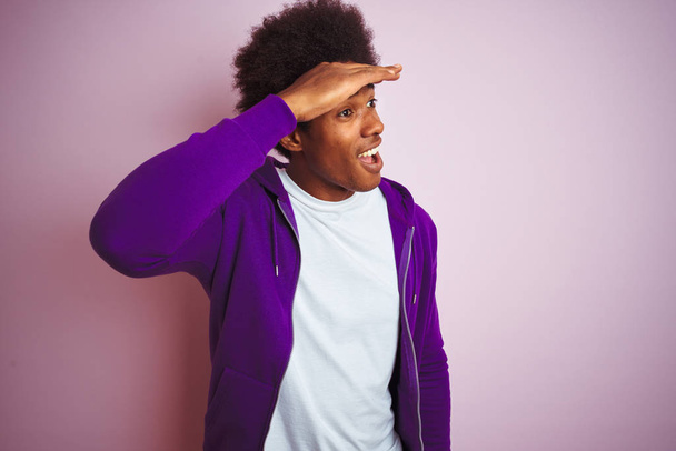 Fiatal afro-amerikai férfi visel lila pulóver állt át elszigetelt rózsaszín háttér nagyon boldog, és mosolyogva keres messze kézzel feje fölött. Keresés a koncepcióban. - Fotó, kép