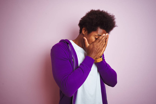孤立したピンクの背景の上に立つ紫色のスウェットシャツを着た若いアフリカ系アメリカ人男性は、泣きながら手で顔を覆う悲しい表情を浮かべていた。うつ病の概念. - 写真・画像