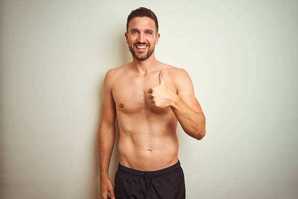 Young handsome shirtless man over isolated background doing happy thumbs up gesture with hand. Approving expression looking at the camera showing success. - Photo, Image