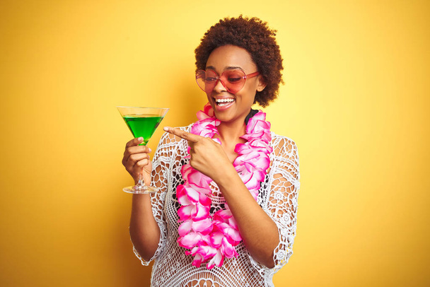 Giovane donna afro americana con capelli afro indossando fiori hawaiani lei e bevendo un cocktail molto felice puntando con mano e dito
 - Foto, immagini