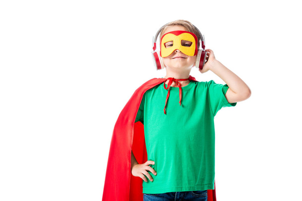 Вид спереди улыбающегося дошкольника в маске и красном плаще героя, слушающего музыку в наушниках и смотрящего в камеру, изолированную от белого
 - Фото, изображение
