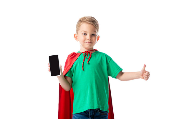 Vooraanzicht van glimlachend kleuters Kid in rode held mantel toont smartphone met leeg scherm en toont duim omhoog geïsoleerd op wit - Foto, afbeelding