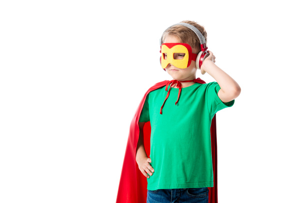 menino pré-escolar em capa de herói vermelho e máscara ouvir música em fones de ouvido e olhando para longe isolado no branco
 - Foto, Imagem