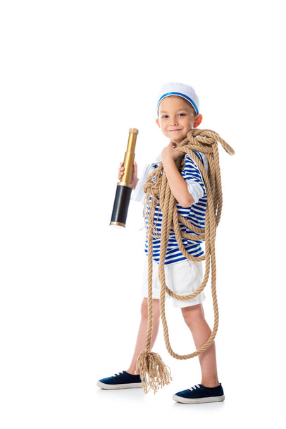 full length view of smiling preschooler child in sailor costume holding spyglass and rope on white - Photo, Image