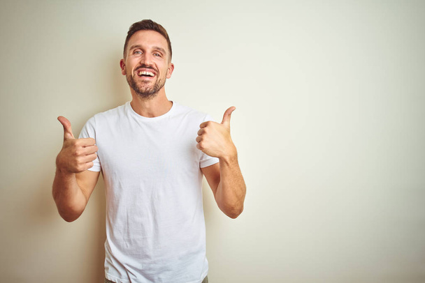 Young handsome man wearing casual white t-shirt over isolated background success sign doing positive gesture with hand, thumbs up smiling and happy. Cheerful expression and winner gesture. - Photo, Image