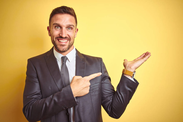 Young handsome business man over yellow isolated background amazed and smiling to the camera while presenting with hand and pointing with finger. - Photo, Image
