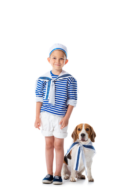 full length view of smiling preschooler child in sailor costume and beagle dog on white - Foto, afbeelding