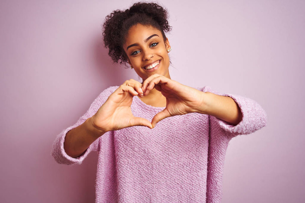 Молоді афро-американської жінки носять зимовий светр стоячи над ізольованою рожевою фоном посміхаючись в коханні робити форму серця символ з рук. Романтична концепція. - Фото, зображення