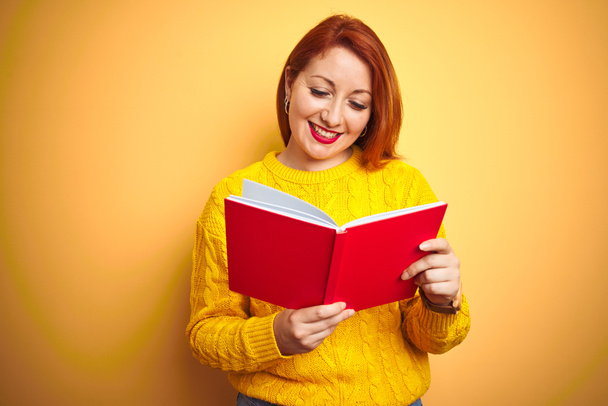 Молодая рыжая учительница читает красную книгу на желтом изолированном фоне со счастливым лицом стоя и улыбаясь с уверенной улыбкой, показывая зубы
 - Фото, изображение
